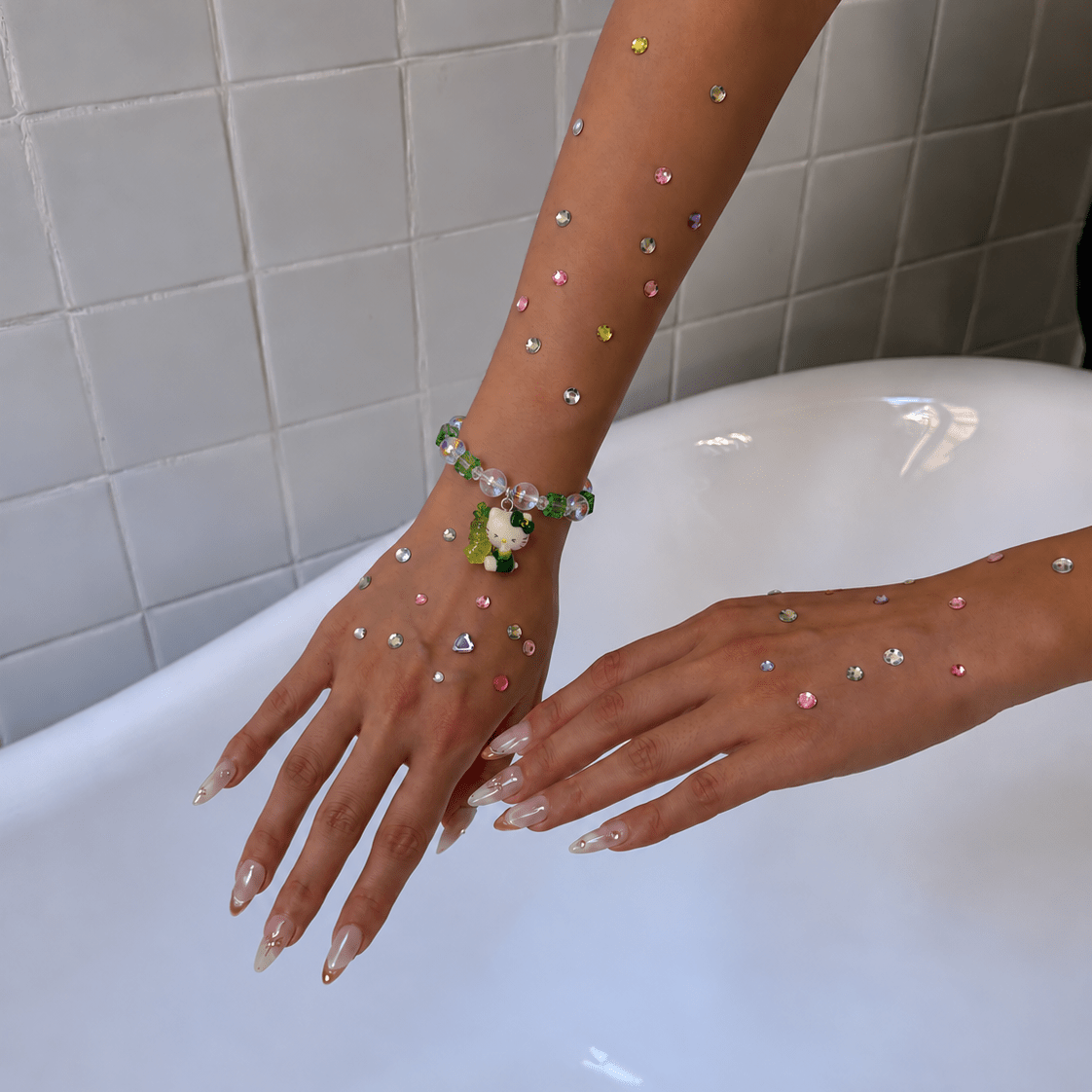 Green and clear crystal bracelet with a small cat charm worn on a wrist decorated with colorful rhinestones.