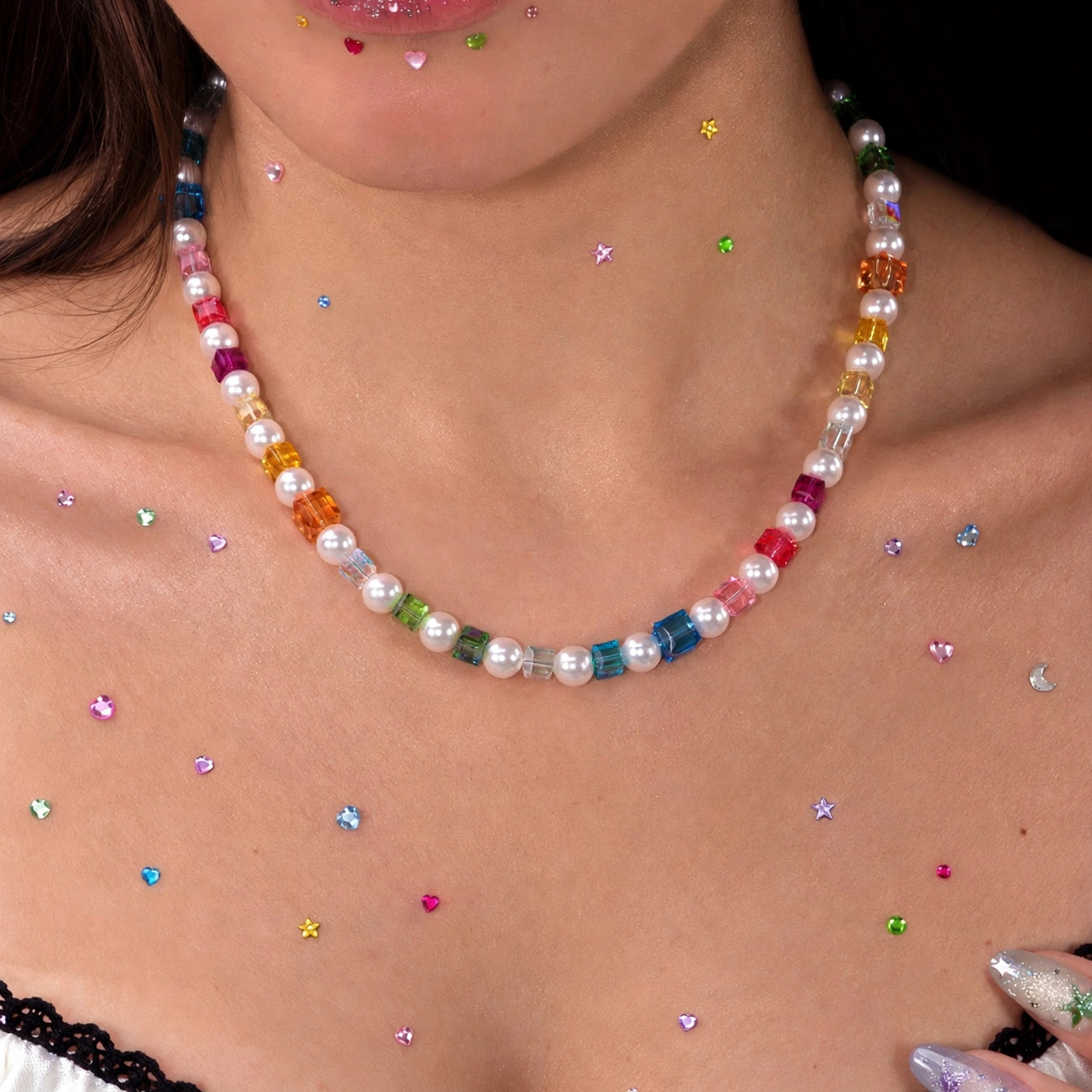 Close-up of a rainbow crystal cube and pearl necklace worn above a white lace-trimmed top, with colorful rhinestones on the skin.