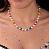Close-up of a rainbow crystal cube and pearl necklace worn above a white lace-trimmed top, with colorful rhinestones on the skin.