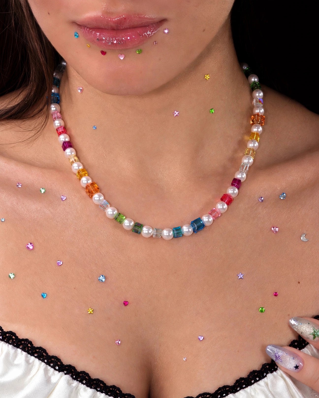 Close-up of a rainbow crystal cube and pearl necklace worn above a white lace-trimmed top, with colorful rhinestones on the skin.