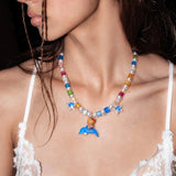 Model wearing a rainbow crystal and white pearl necklace with blue stars and a cat-and-dolphin charm over a white lace top.