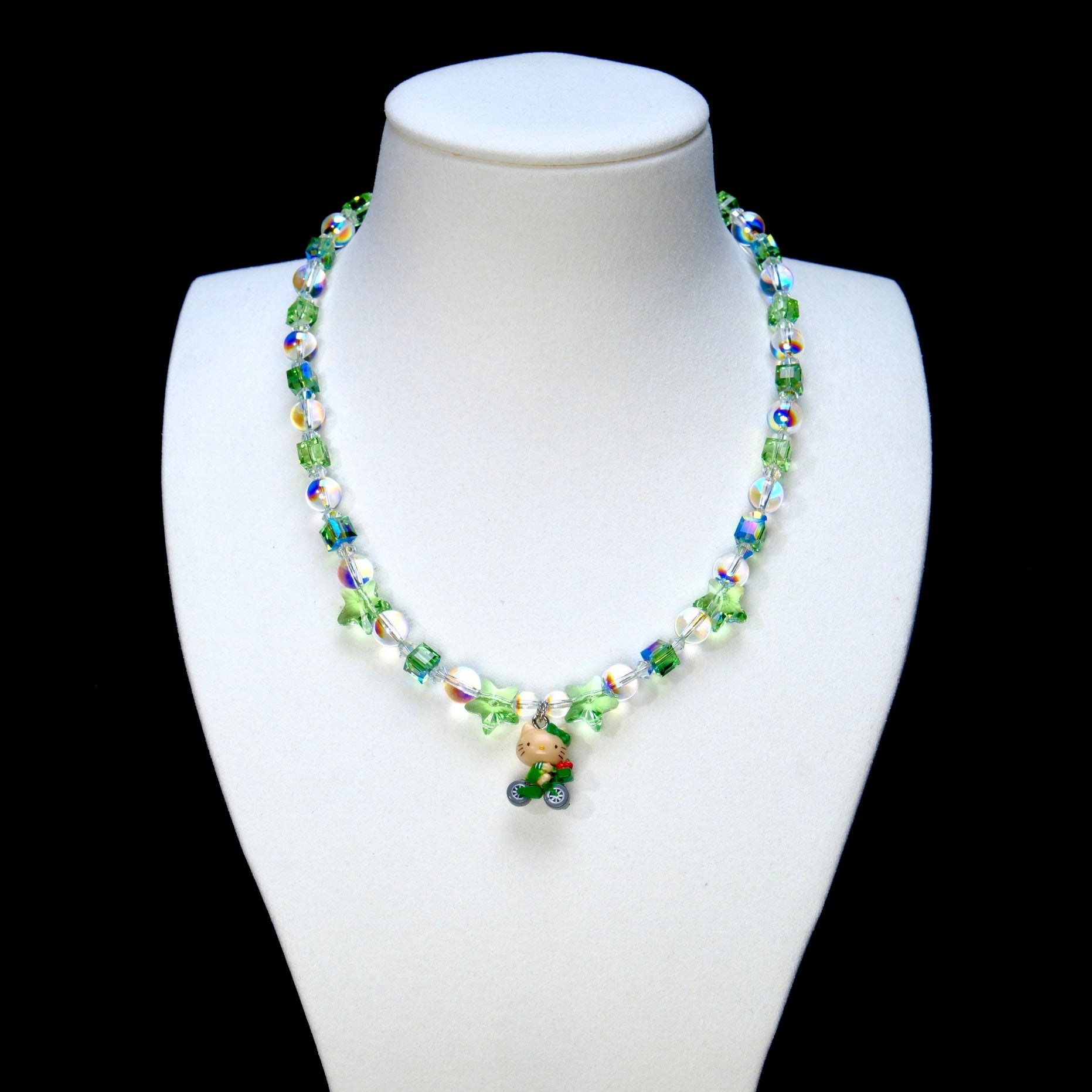 Green and iridescent crystal necklace with green star beads and a bicycle-riding cat charm on a white display bust.