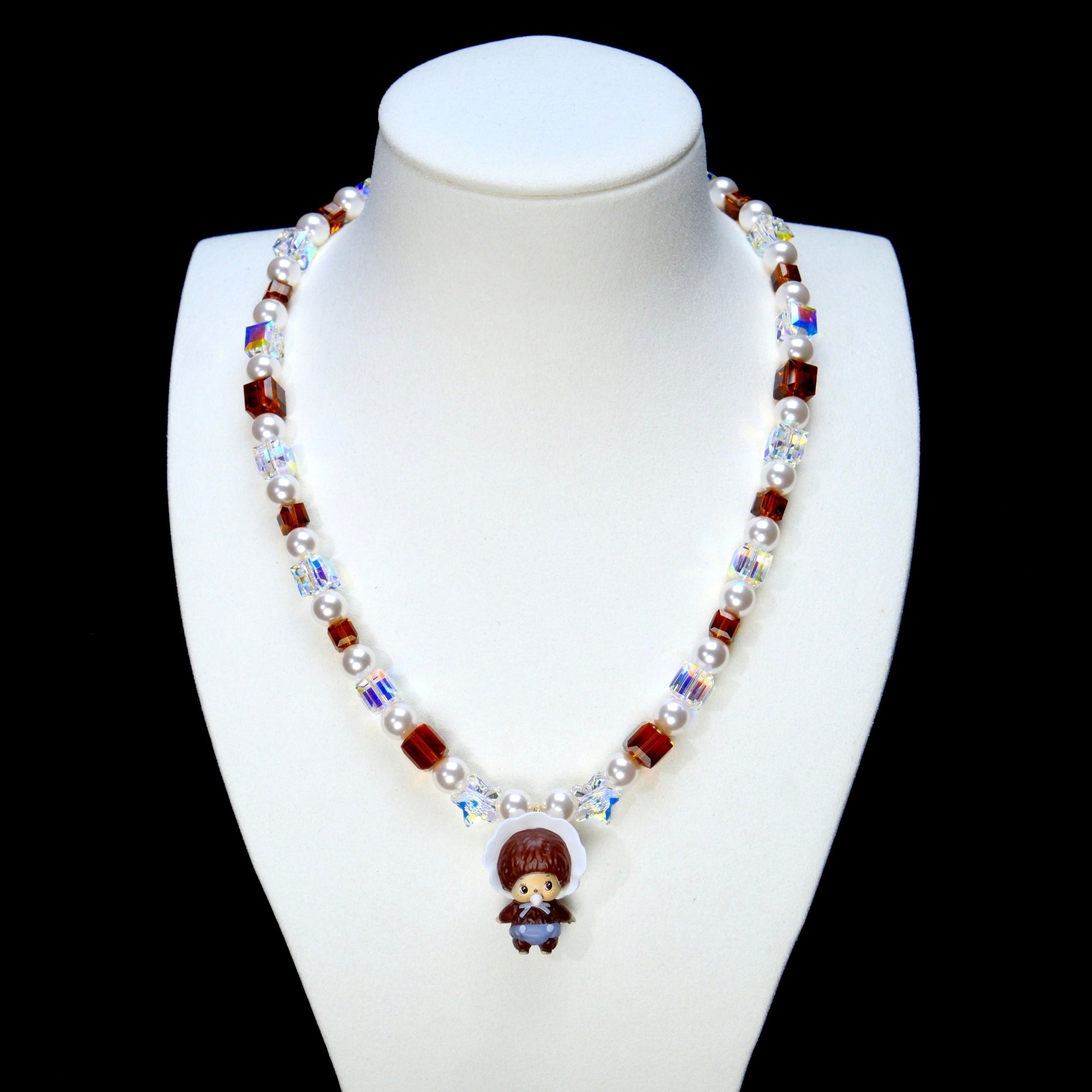 Amber-brown crystal and pearl necklace with iridescent star beads and a bonnet-wearing doll charm on a white display bust.