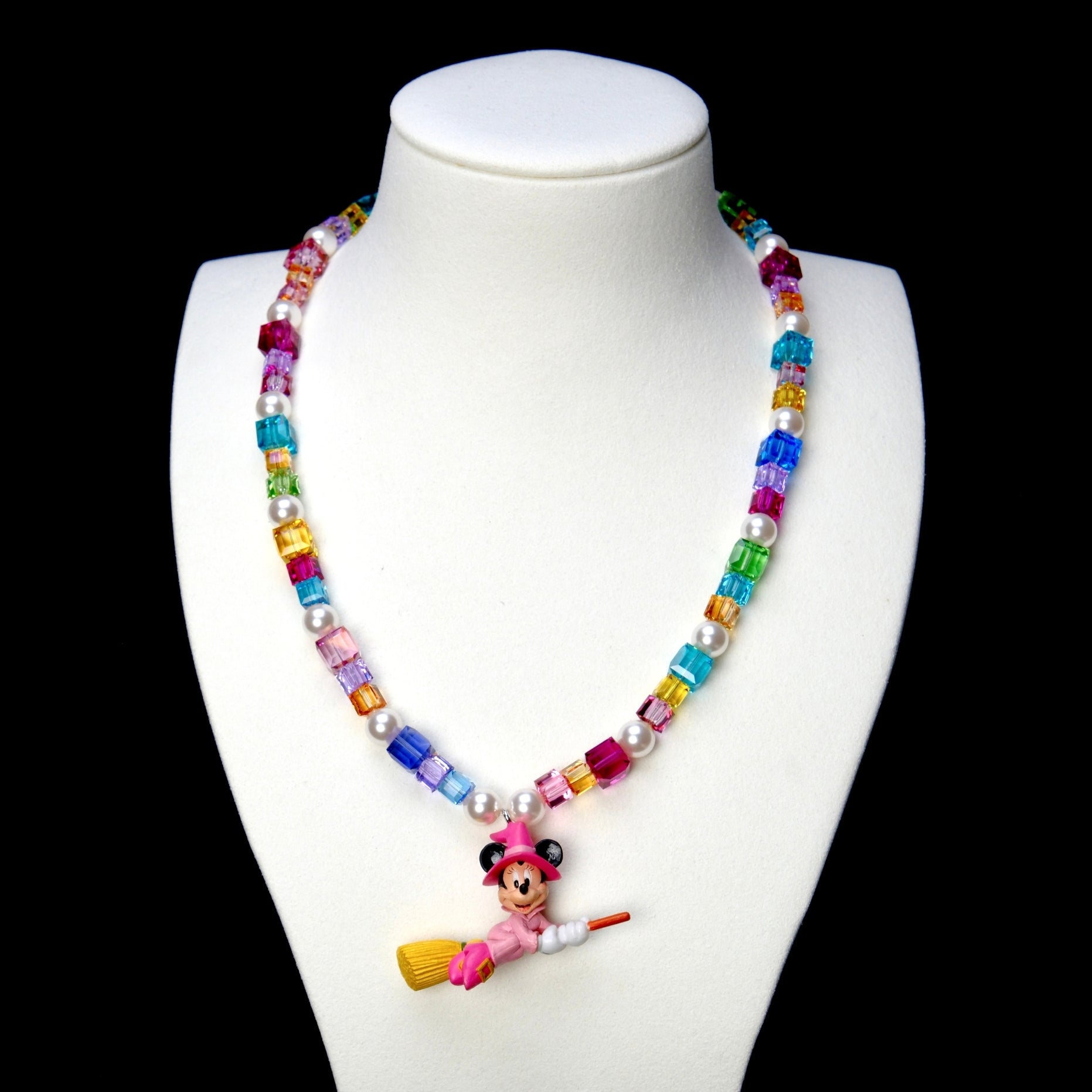 Rainbow crystal and white pearl necklace with a pink witch mouse riding a broom, displayed on a white bust against black.