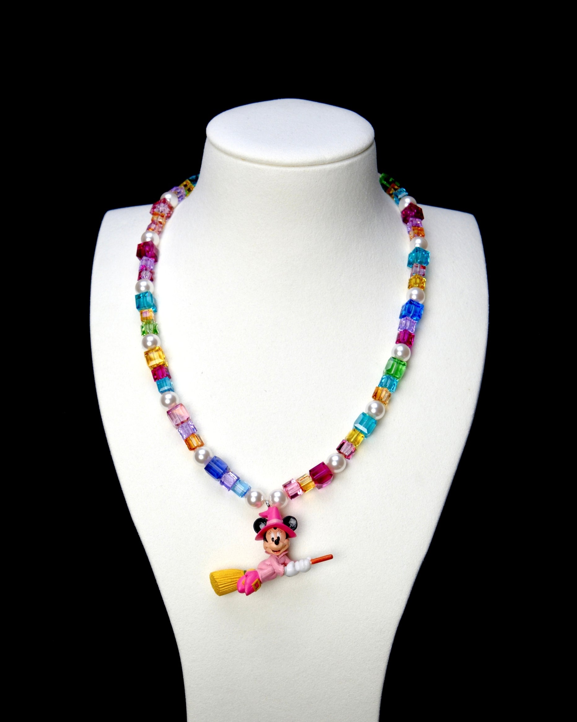 Rainbow crystal and white pearl necklace with a pink witch mouse riding a broom, displayed on a white bust against black.