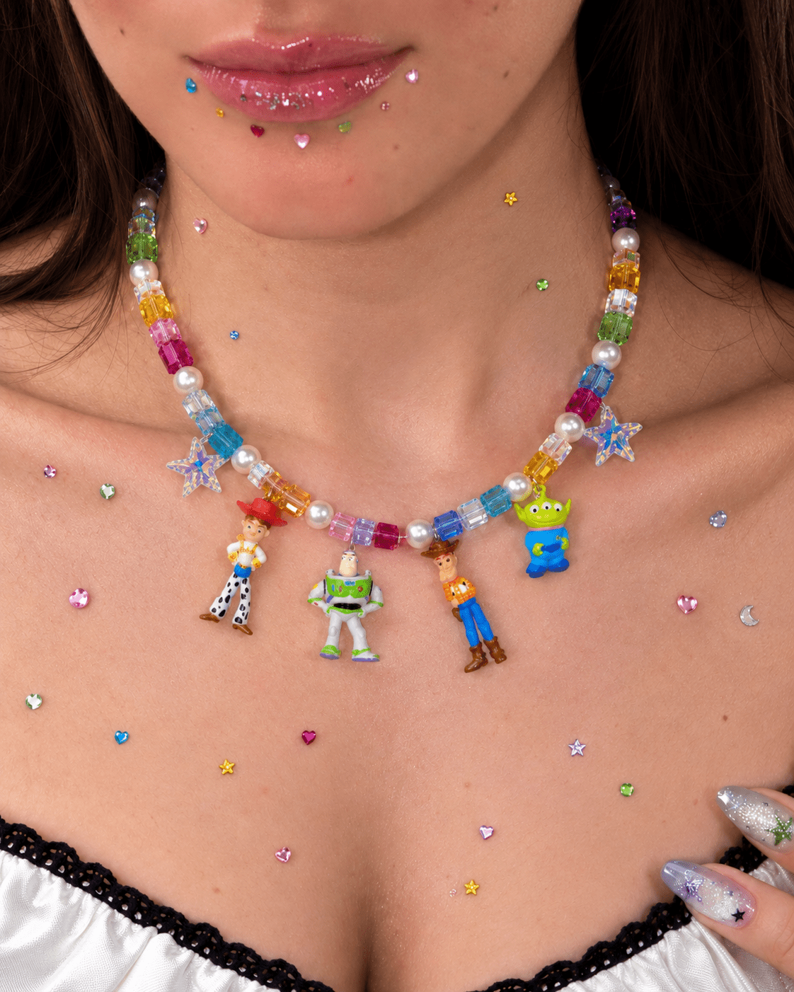 Close-up of a rainbow crystal and pearl necklace with four colorful toy figure charms, worn above a white lace-trimmed top.