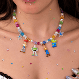 Close-up of a rainbow crystal and pearl necklace with four colorful toy figure charms, worn above a white lace-trimmed top.