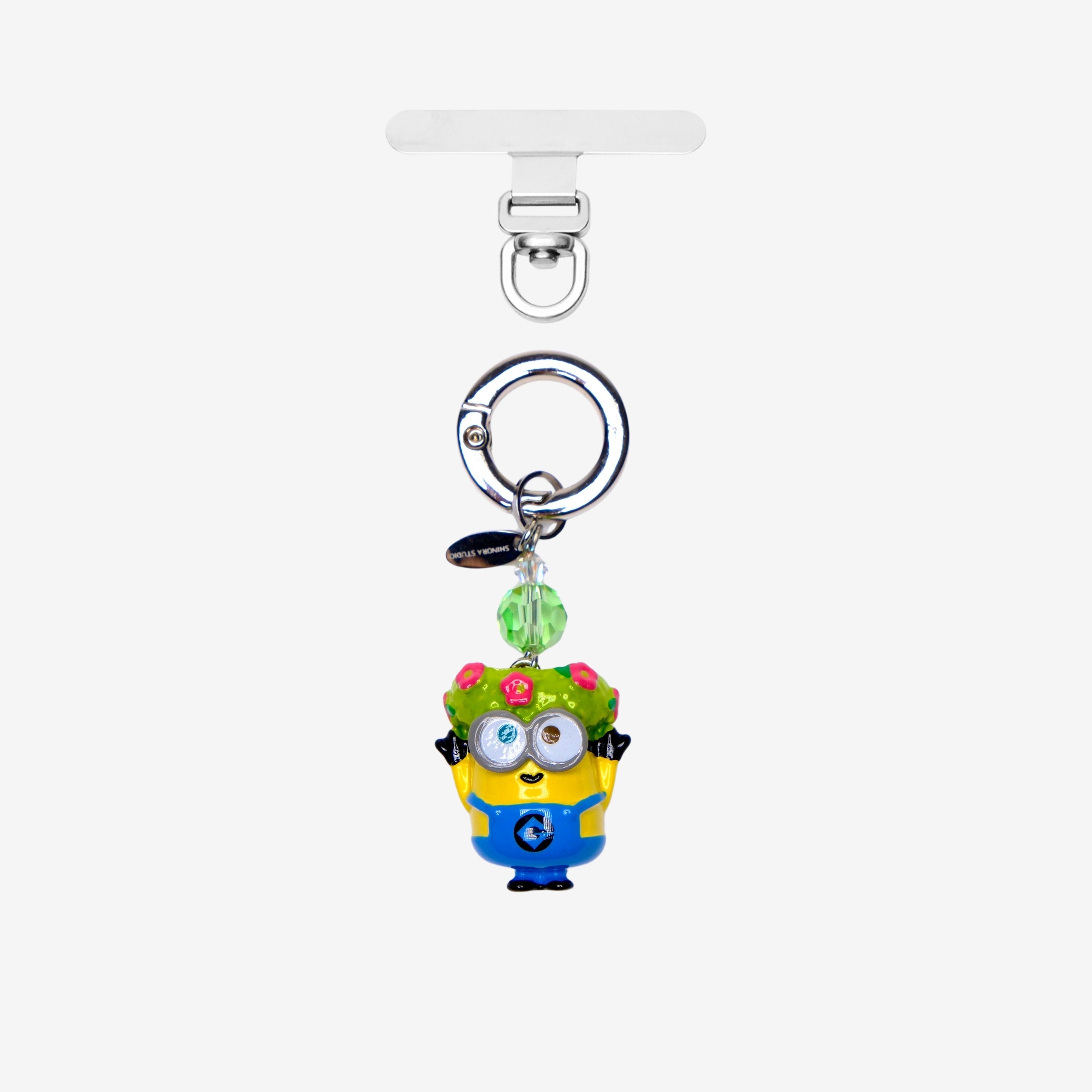 Yellow goggle-eyed character phone charm wearing a floral crown, with a green crystal accent and silver attachment ring.