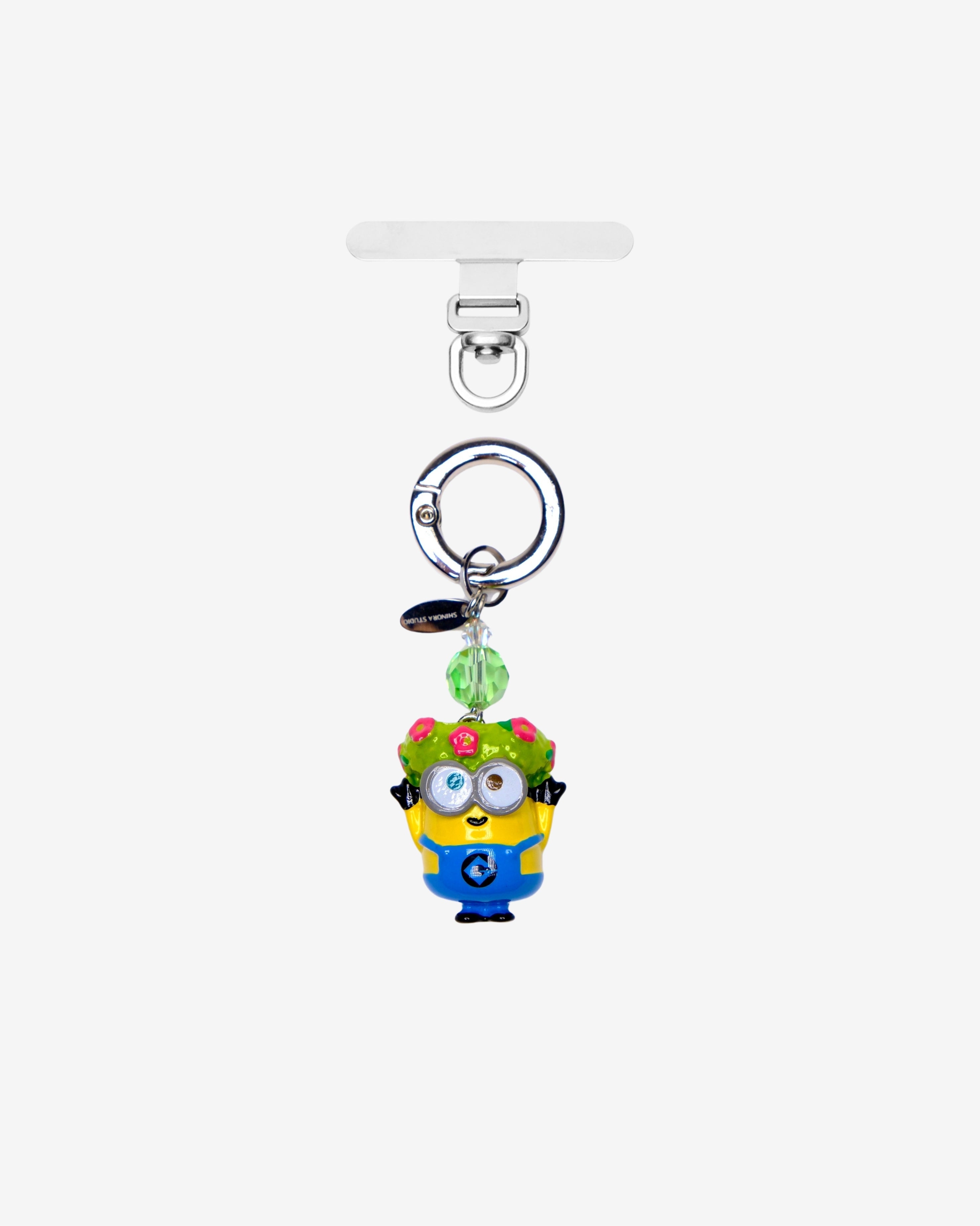Yellow goggle-eyed character phone charm wearing a floral crown, with a green crystal accent and silver attachment ring.