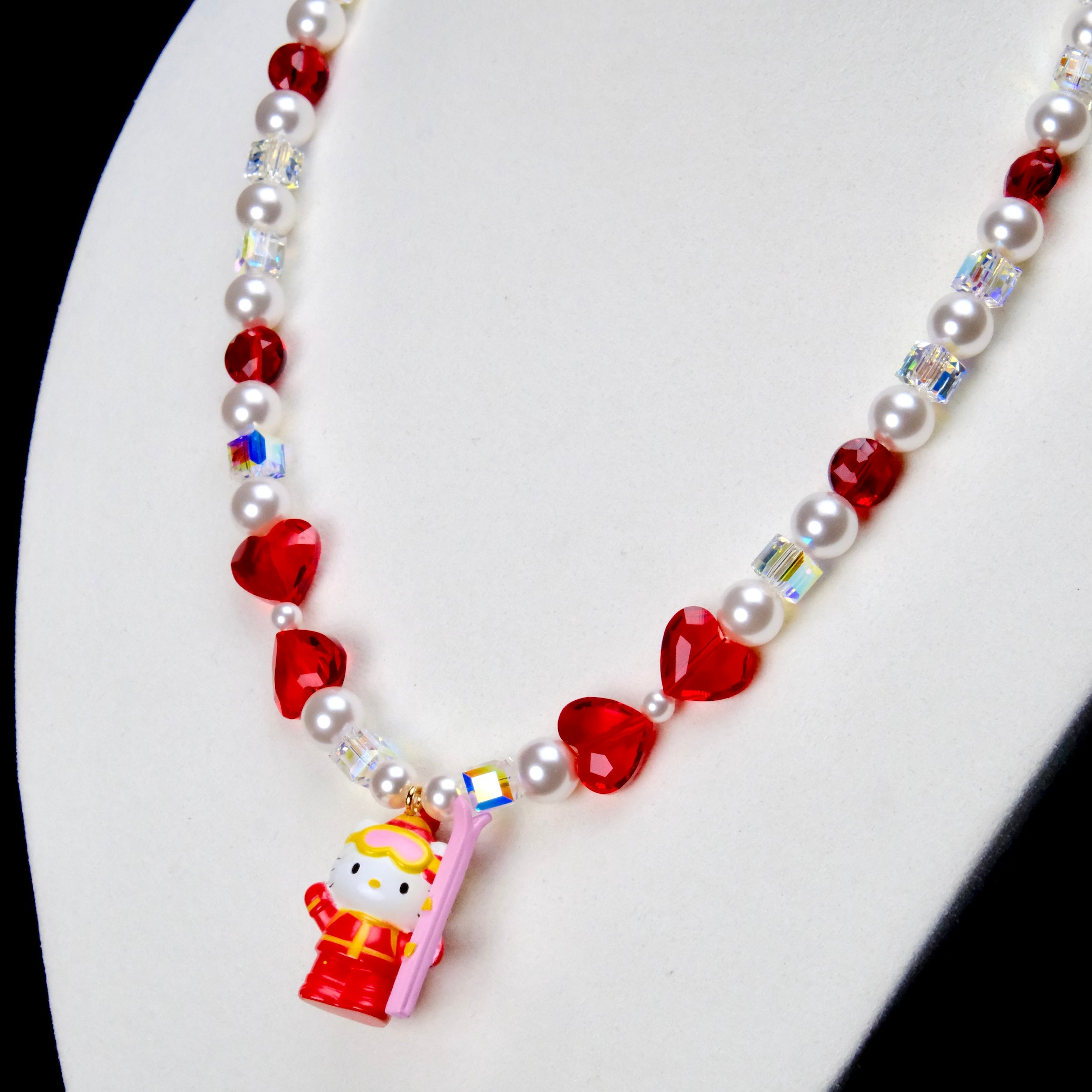 Angled close-up of Ruby Heart Snow Day Charm Necklace showing red heart crystals, white pearls and a red winter-outfit cat pendant.