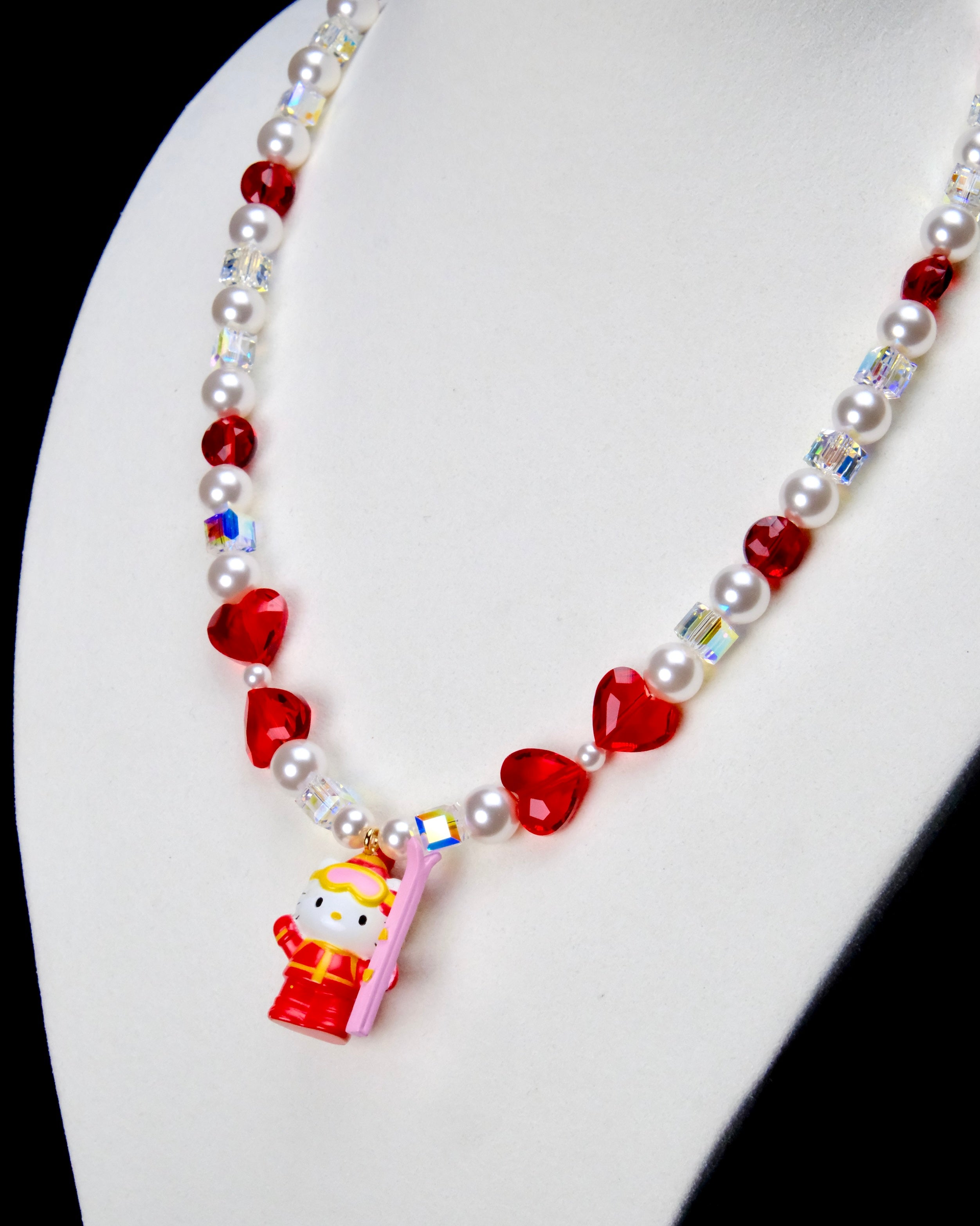 Angled close-up of Ruby Heart Snow Day Charm Necklace showing red heart crystals, white pearls and a red winter-outfit cat pendant.
