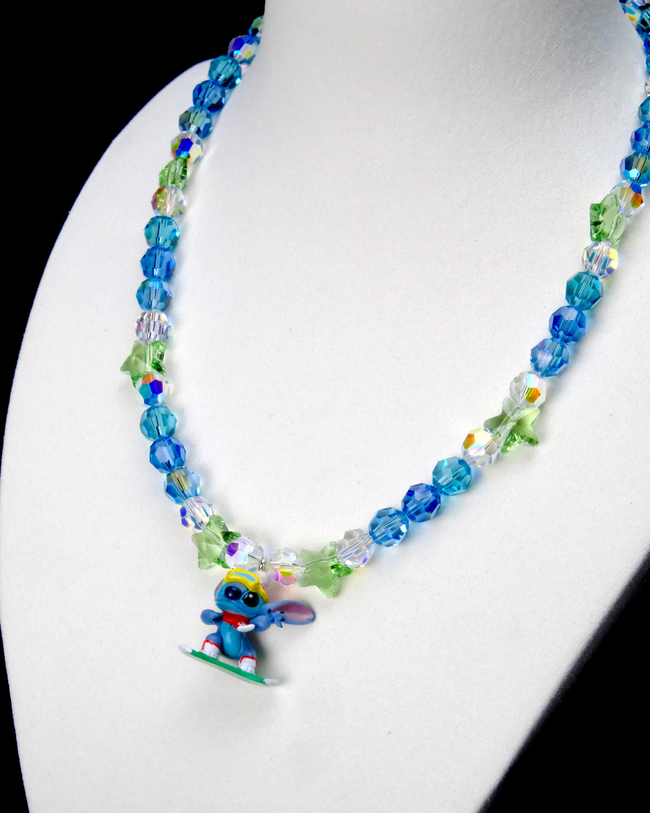 Angled close-up of Ocean Blue Star Rider Charm Necklace showing blue crystals, green star beads and a blue character on a green board.
