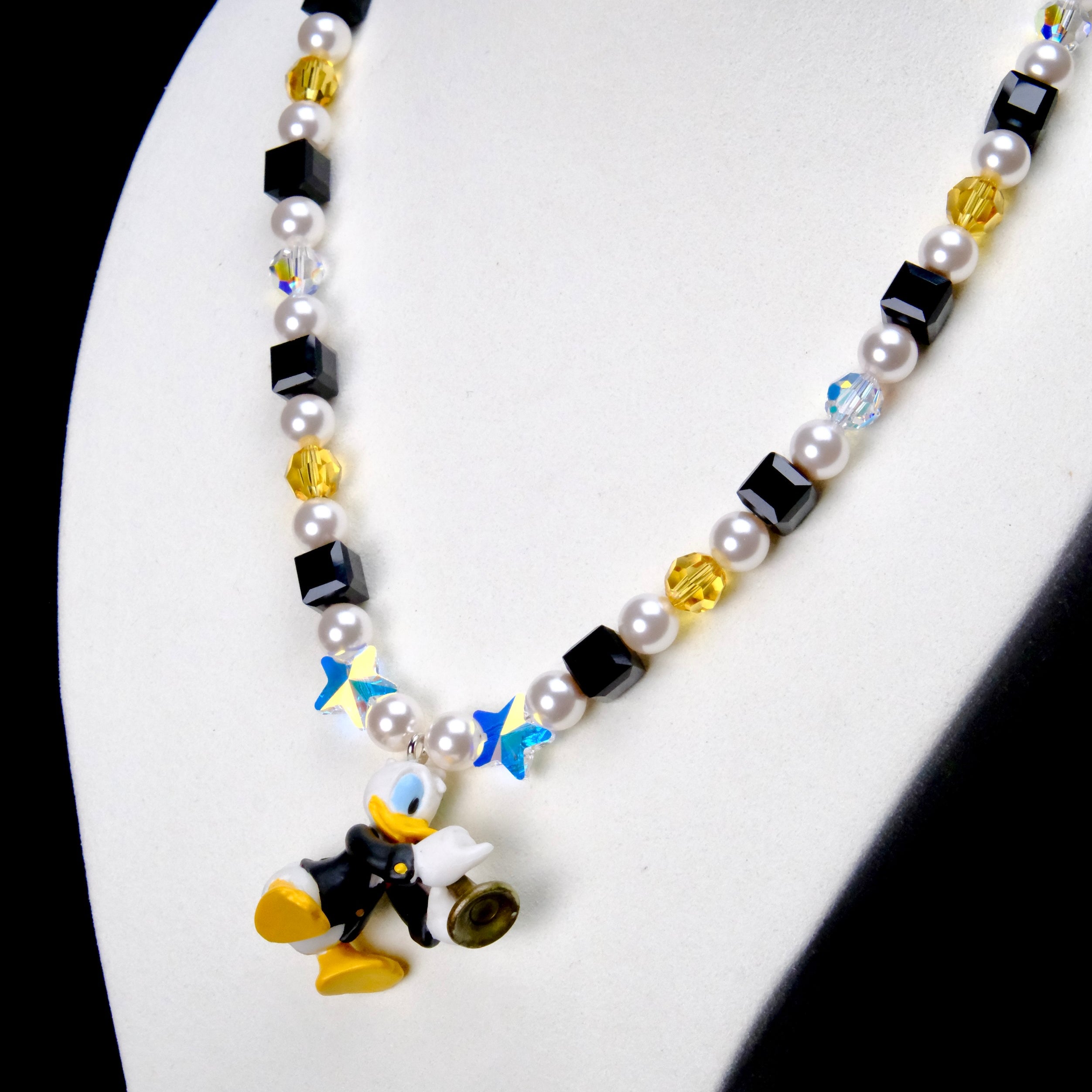 Angled close-up of Black & Gold Melody Charm Necklace showing black and yellow crystals, white pearls and a duck with a brass instrument.