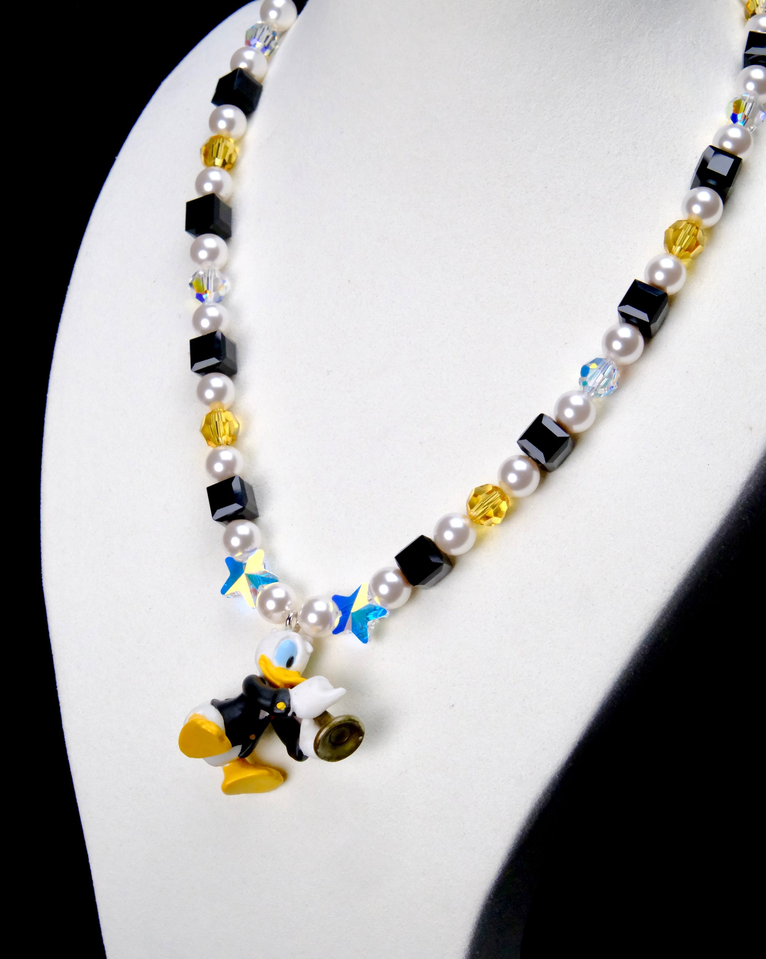 Angled close-up of Black & Gold Melody Charm Necklace showing black and yellow crystals, white pearls and a duck with a brass instrument.