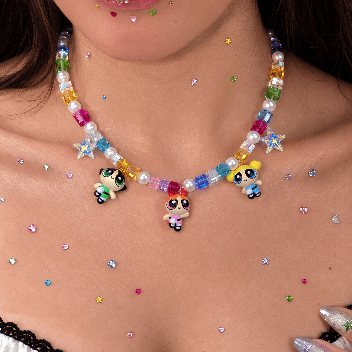 Close-up of a rainbow crystal and pearl necklace with three cartoon girl charms, worn above a white lace-trimmed top.