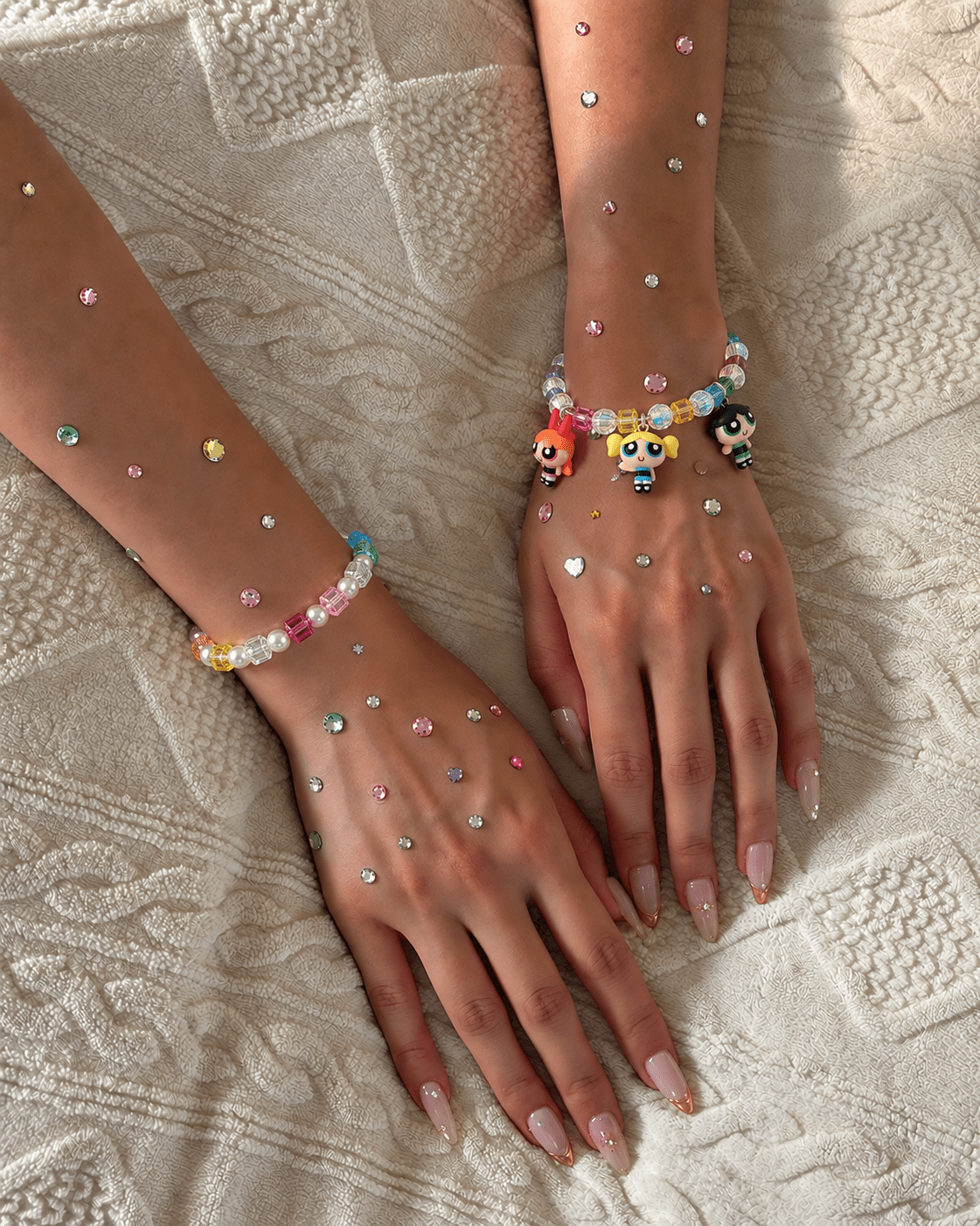 Two gem-decorated wrists wearing rainbow crystal bracelets, with three colorful character charms on one bracelet.