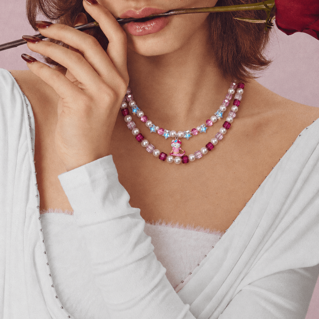 Pink crystal and pearl double-layer necklace with a small skiing cat charm, worn with a white top while holding a rose.