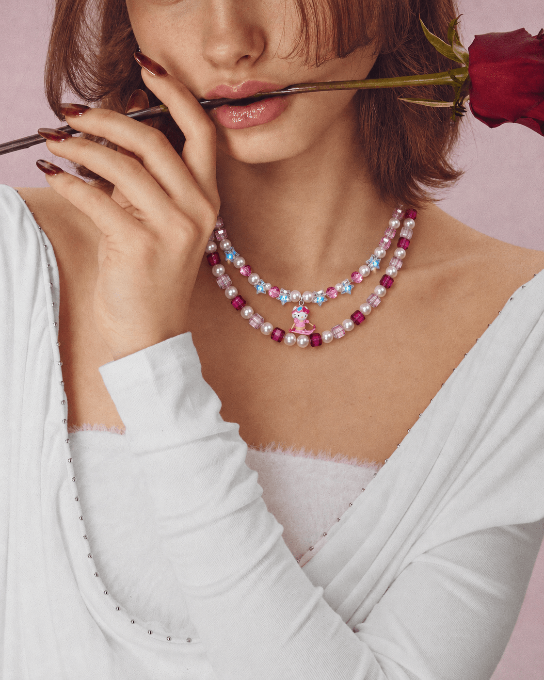 Pink crystal and pearl double-layer necklace with a small skiing cat charm, worn with a white top while holding a rose.