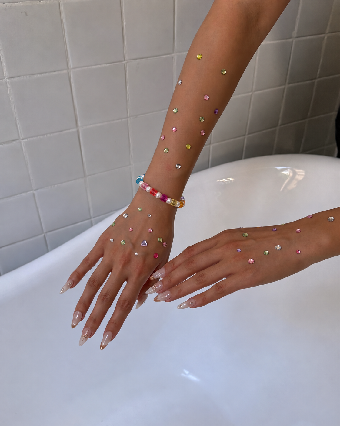Rainbow crystal cube and pearl bracelet worn on a wrist above a bathtub, with colorful rhinestones decorating the hands.