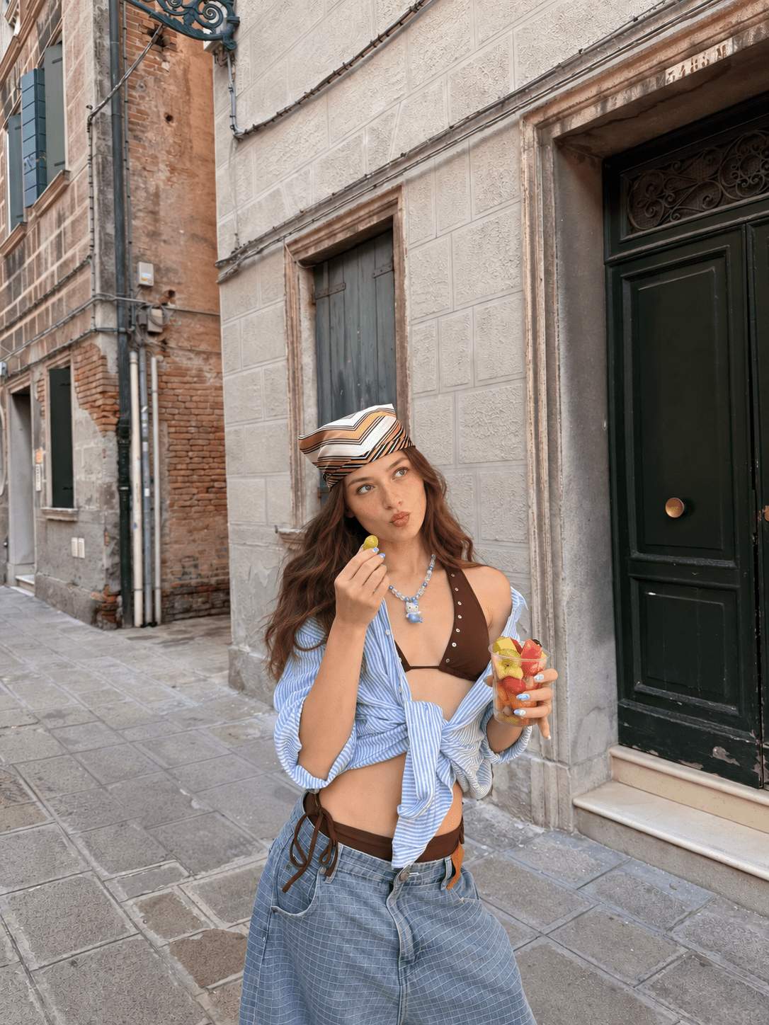 Model wearing a blue pearl and crystal mermaid cat necklace with a striped shirt, holding a cup of fruit on a stone street.