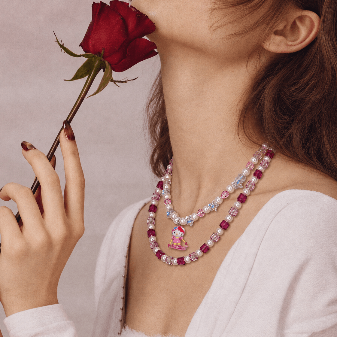 Side view of a pink crystal and pearl double-layer necklace with a skiing cat charm, worn with a white top beside a red rose.