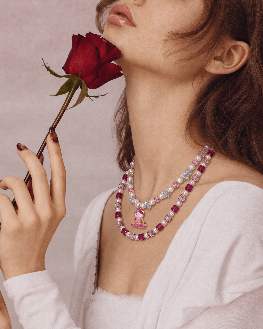 Side view of a pink crystal and pearl double-layer necklace with a skiing cat charm, worn with a white top beside a red rose.