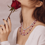Side view of a pink crystal and pearl double-layer necklace with a skiing cat charm, worn with a white top beside a red rose.