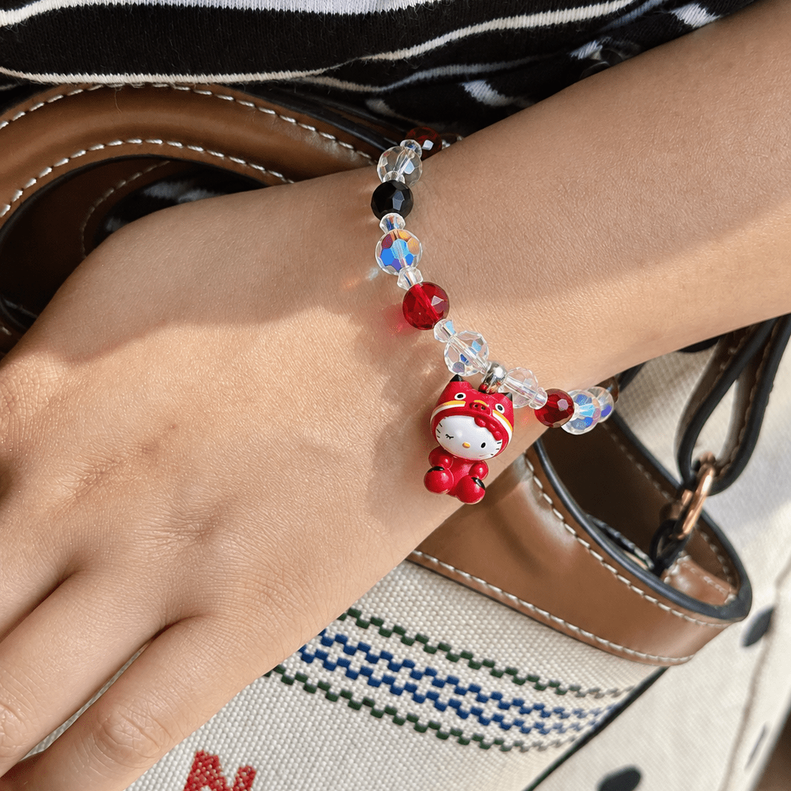 Ruby Costume Cat Charm Bracelet with red, black and iridescent beads and a red-costume cat charm, worn above a cream woven handbag.