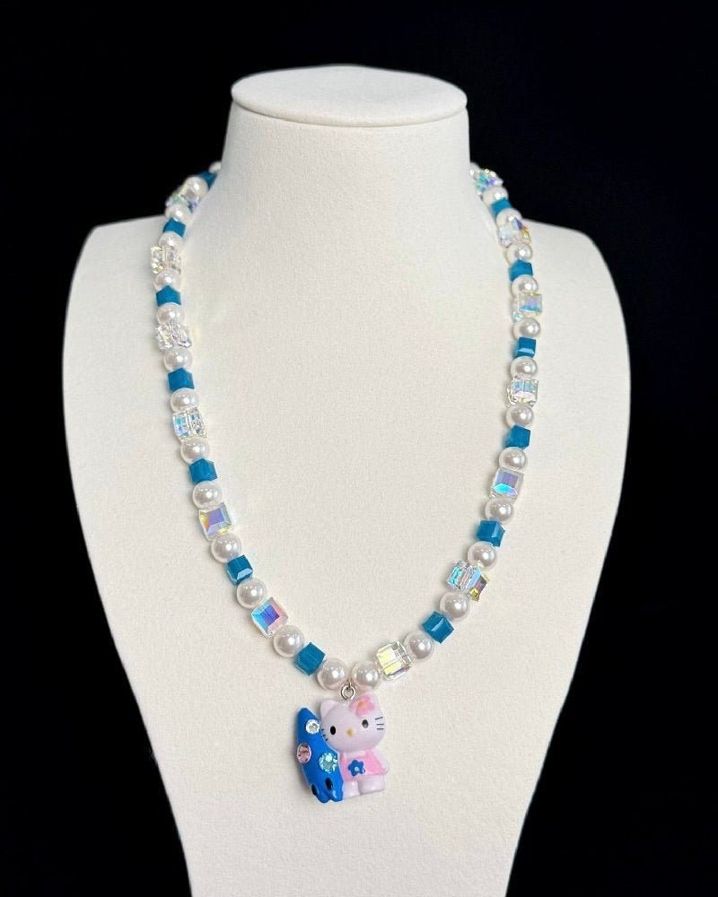 Front view of a blue crystal and pearl necklace with a colorful cat charm on a white jewelry bust against a black background.