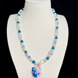 Front view of a blue crystal and pearl necklace with a colorful cat charm on a white jewelry bust against a black background.