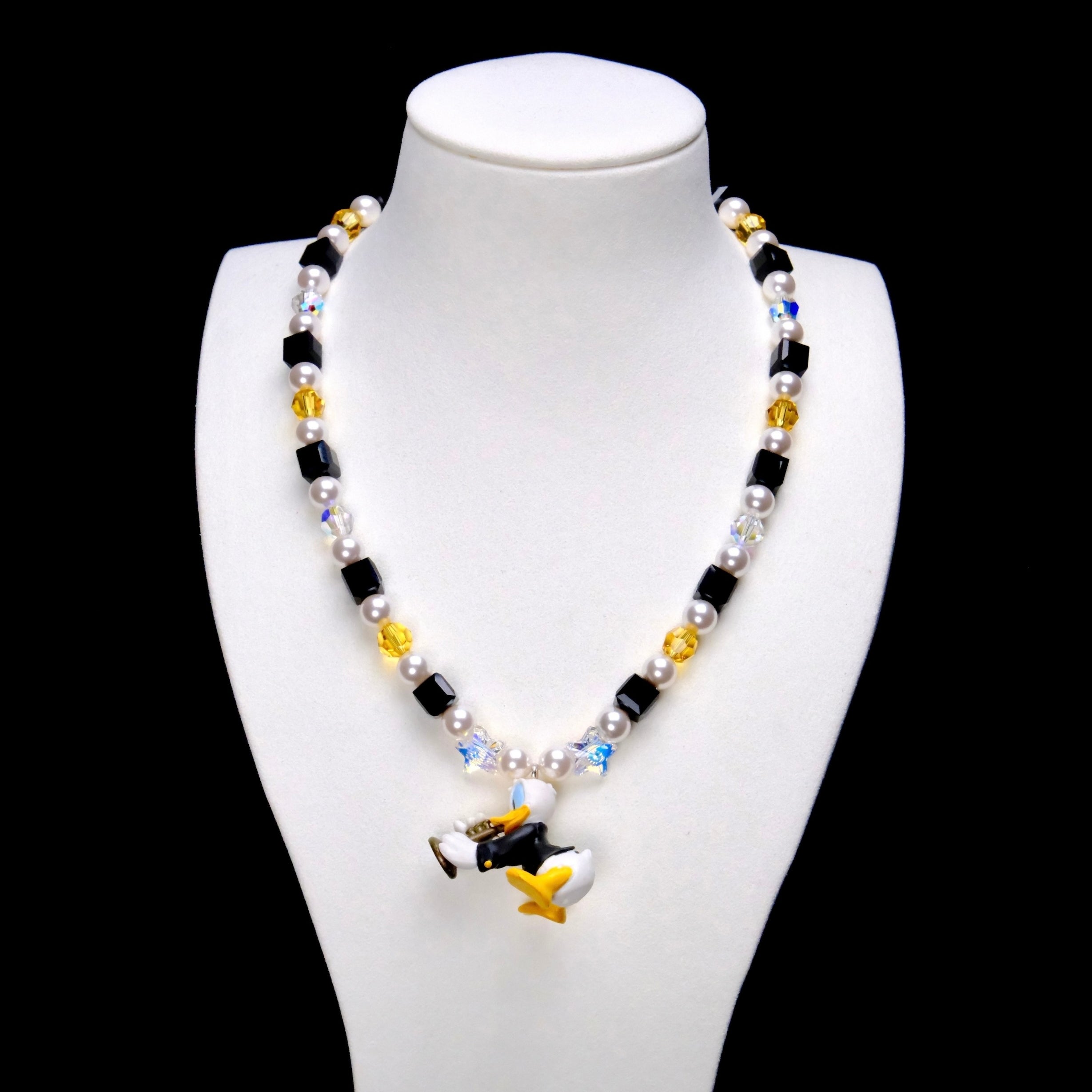Black crystal cubes, yellow crystal rounds and white pearl beads frame a Donald Duck charm in a dark outfit, holding a gold-colored musical instrument.
