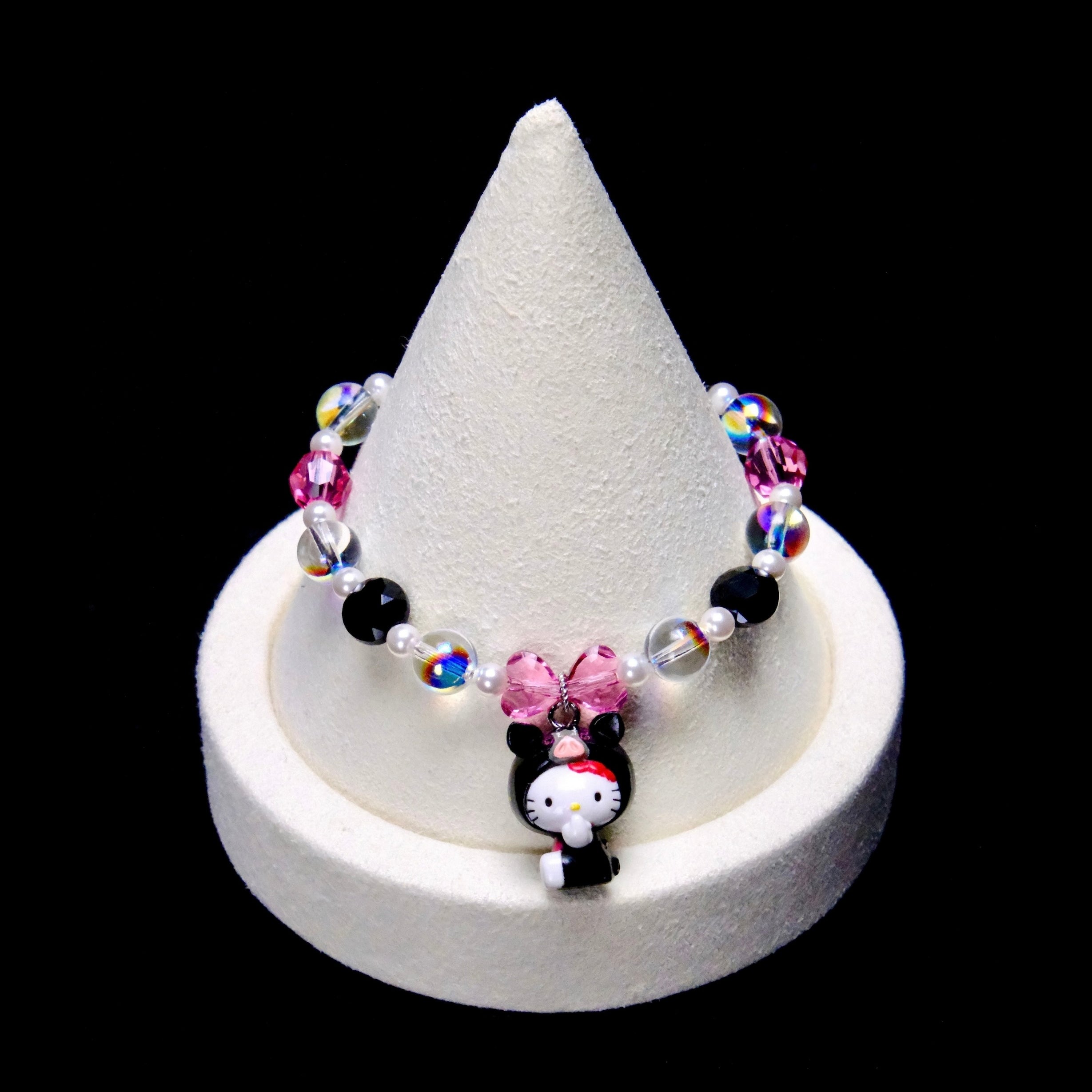 Pink, black and clear iridescent crystal rounds with small white pearl beads frame a pink crystal bow and a Hello Kitty charm wearing a black animal hood with a pink snout.