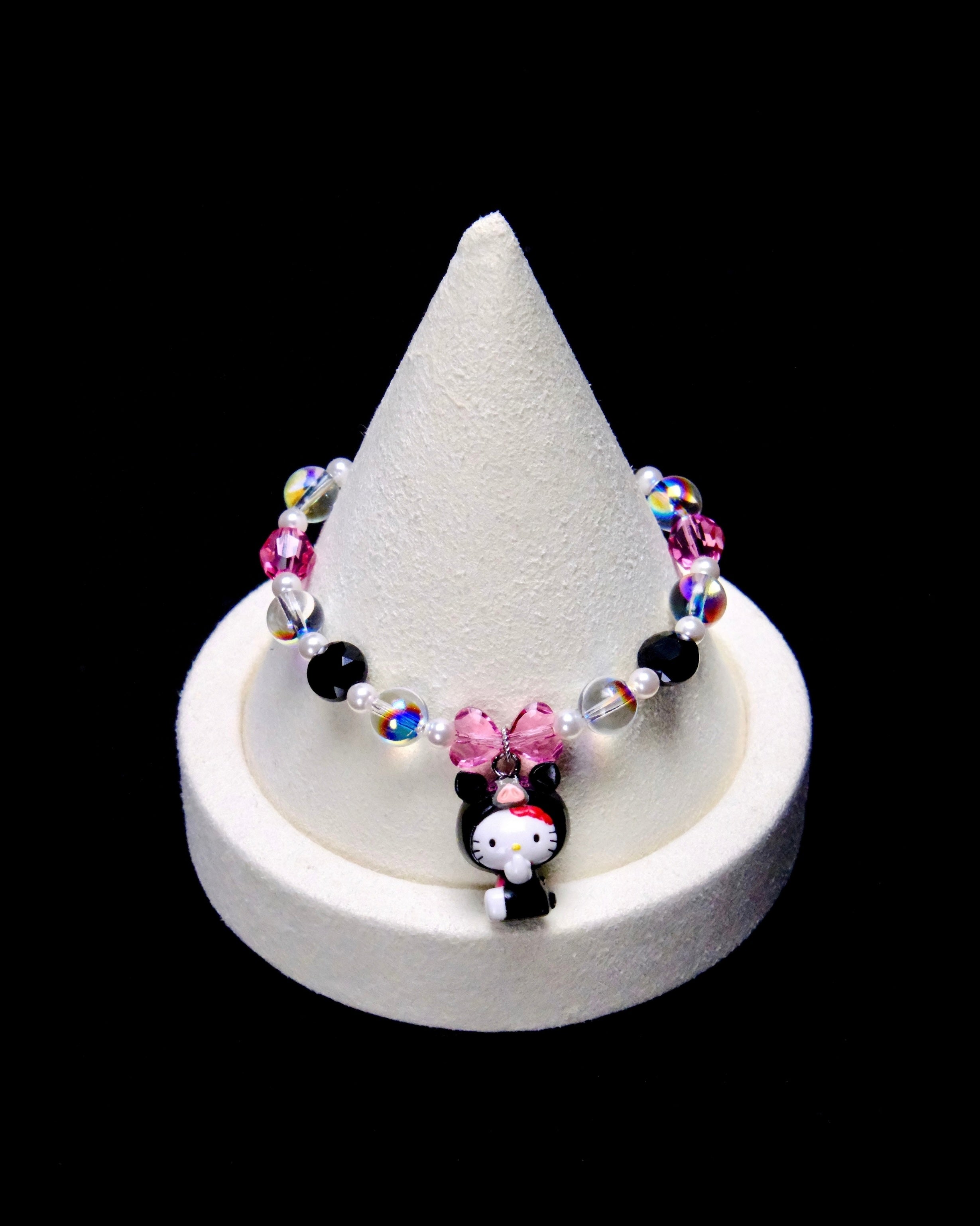 Pink, black and clear iridescent crystal rounds with small white pearl beads frame a pink crystal bow and a Hello Kitty charm wearing a black animal hood with a pink snout.