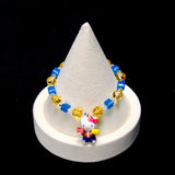 Bright blue crystal cubes and golden-yellow crystal rounds surround a Hello Kitty charm wearing an orange jacket and blue trousers, holding colorful accessories.