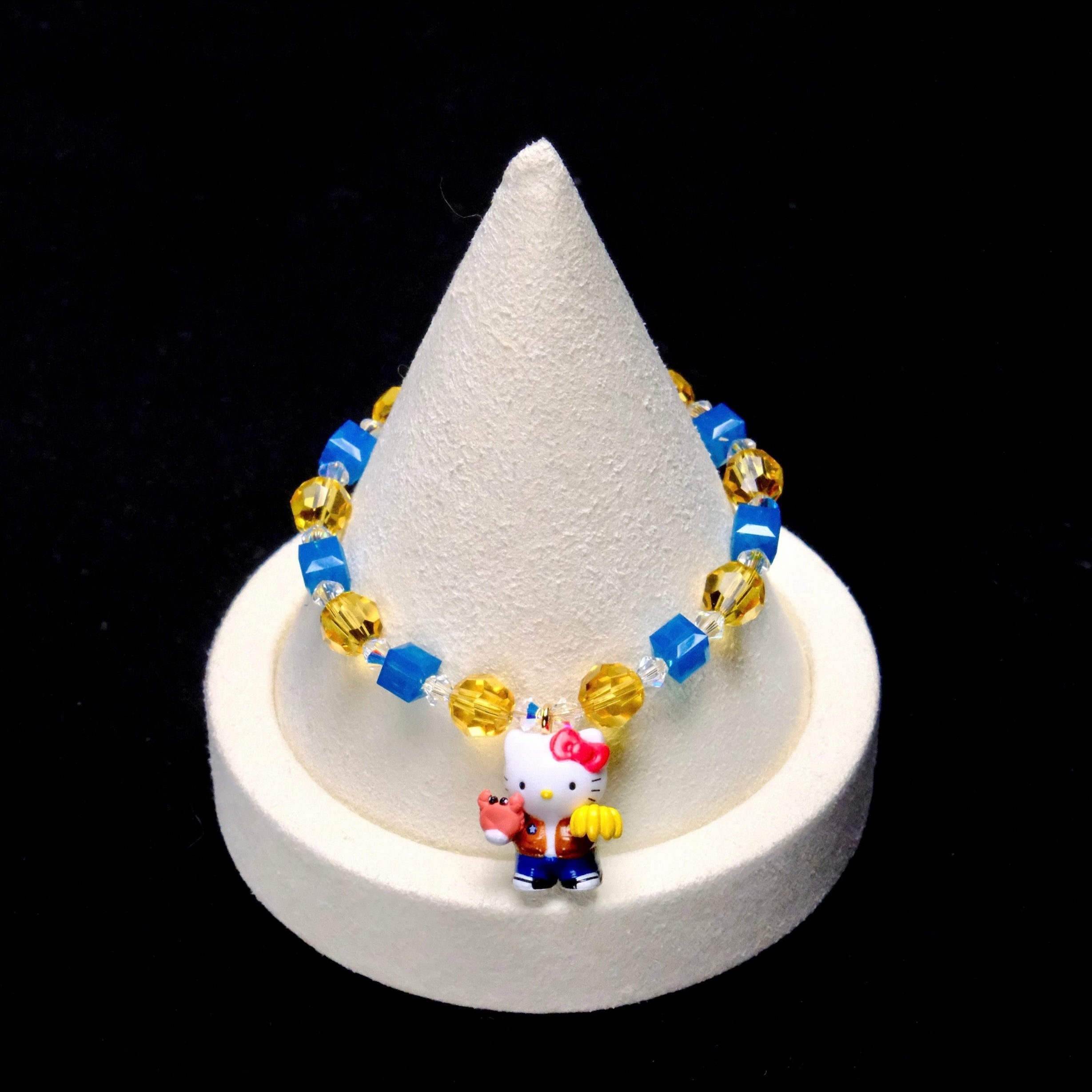 Bright blue crystal cubes and golden-yellow crystal rounds surround a Hello Kitty charm wearing an orange jacket and blue trousers, holding colorful accessories.