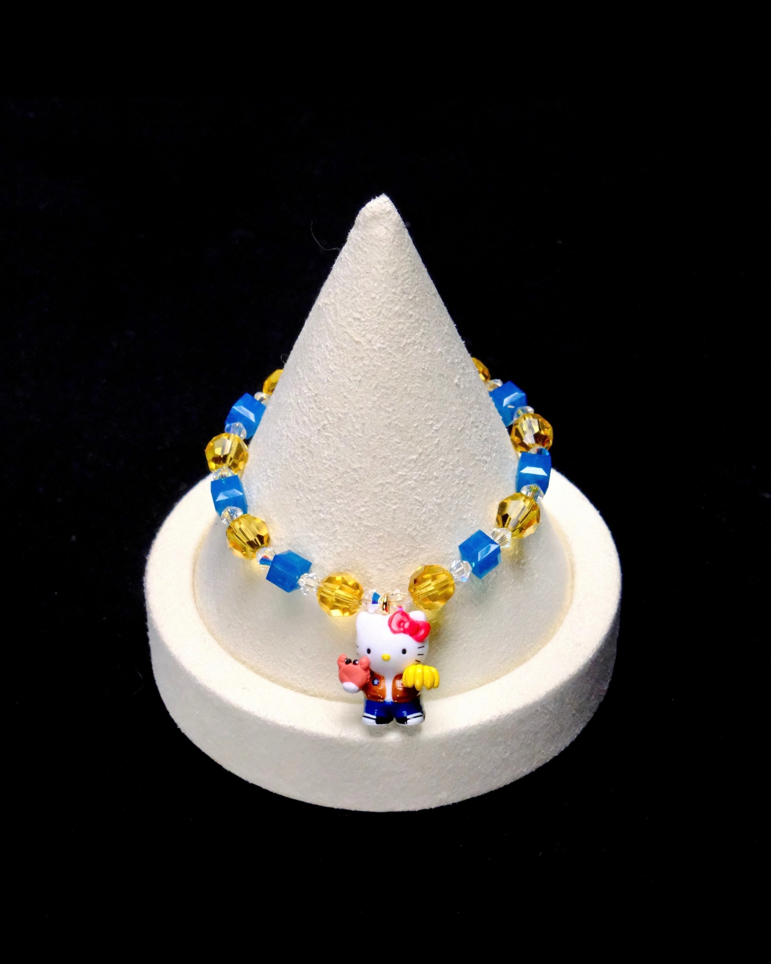 Bright blue crystal cubes and golden-yellow crystal rounds surround a Hello Kitty charm wearing an orange jacket and blue trousers, holding colorful accessories.