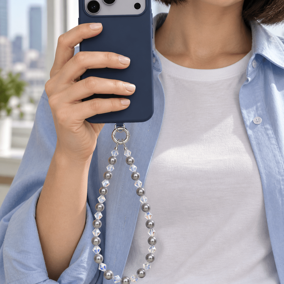 Woman in a pale blue shirt holding a navy phone with a gray pearl and clear crystal phone chain.