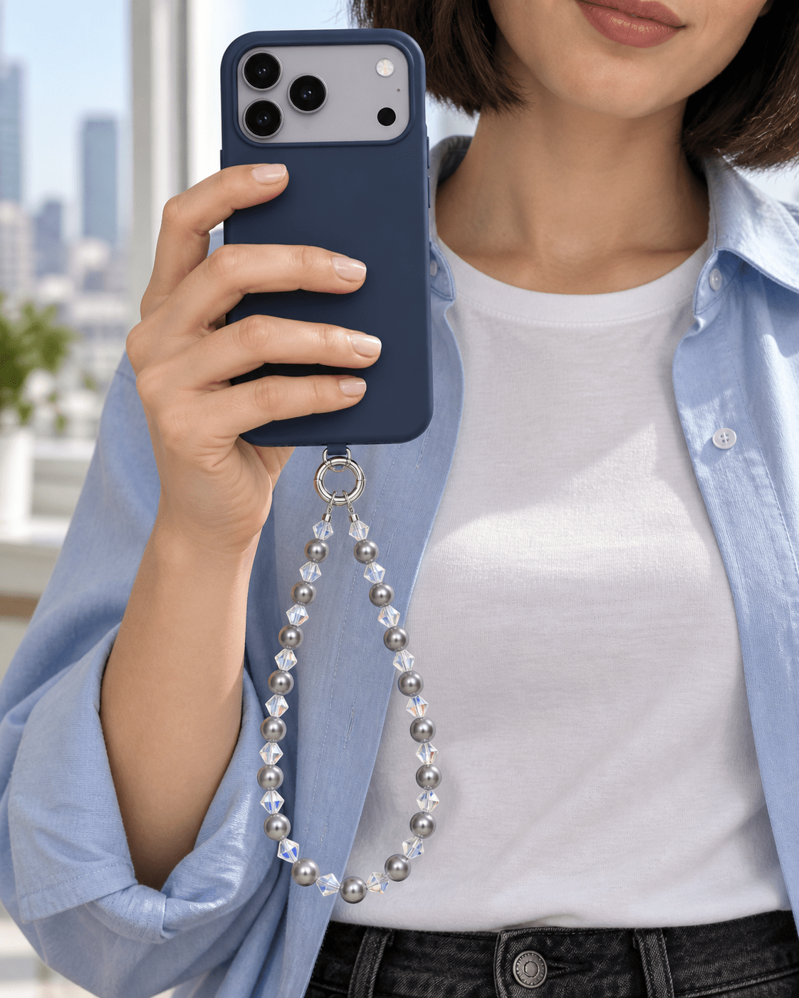 Woman in a pale blue shirt holding a navy phone with a gray pearl and clear crystal phone chain.