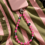 Pink crystal phone chain with a black heart and black cubes, attached to a pink phone on olive and pink striped fabric.
