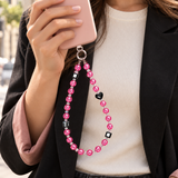 Woman in a black blazer holding a pink phone with a pink crystal phone chain and black heart detail.