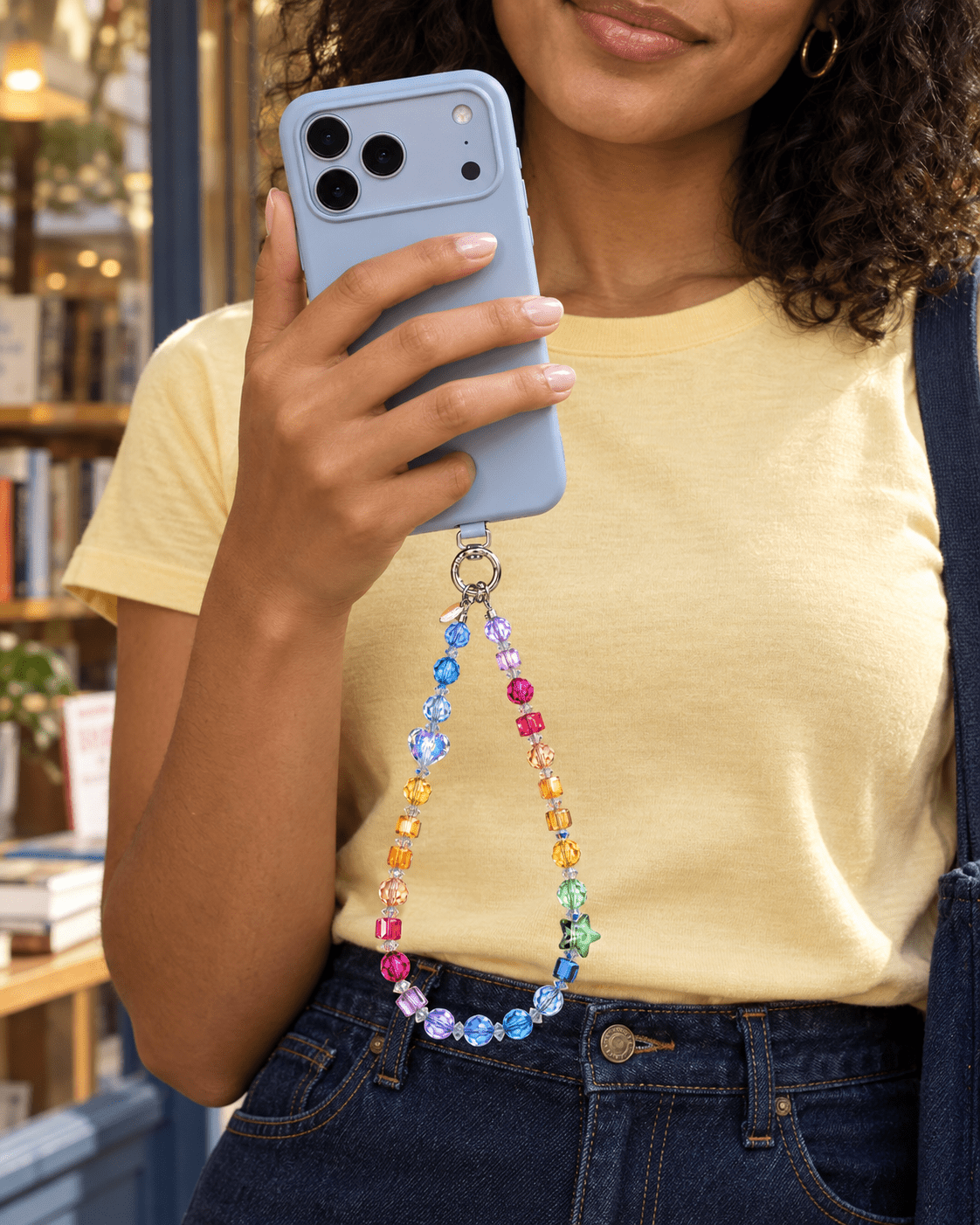 Woman in a yellow T-shirt holding a blue phone with a rainbow crystal phone chain.