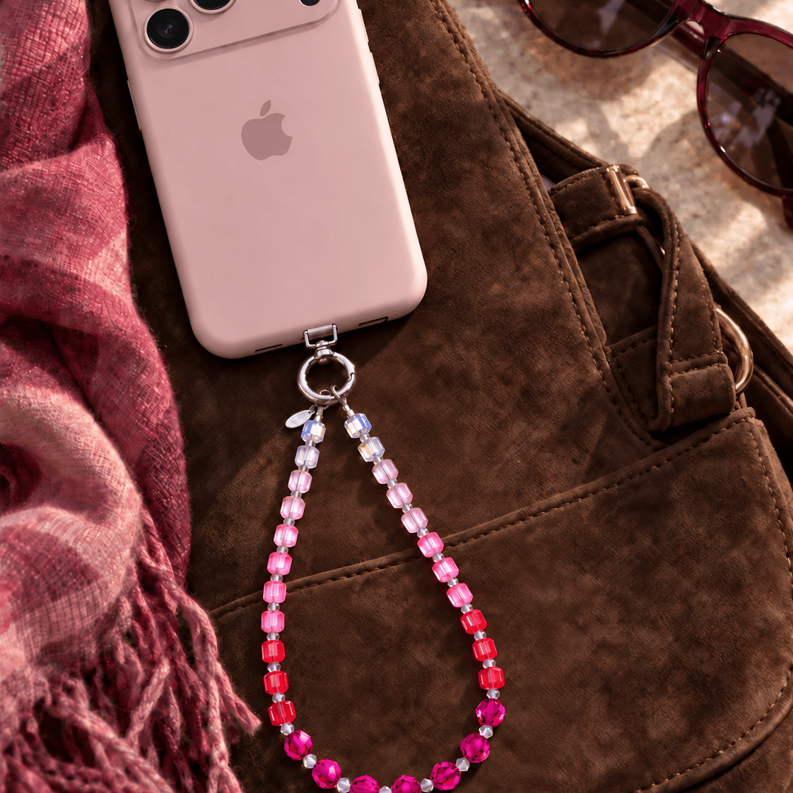 Rose ombre crystal phone chain with pale pink, pink, red and plum beads, attached to a pink phone on a brown bag.