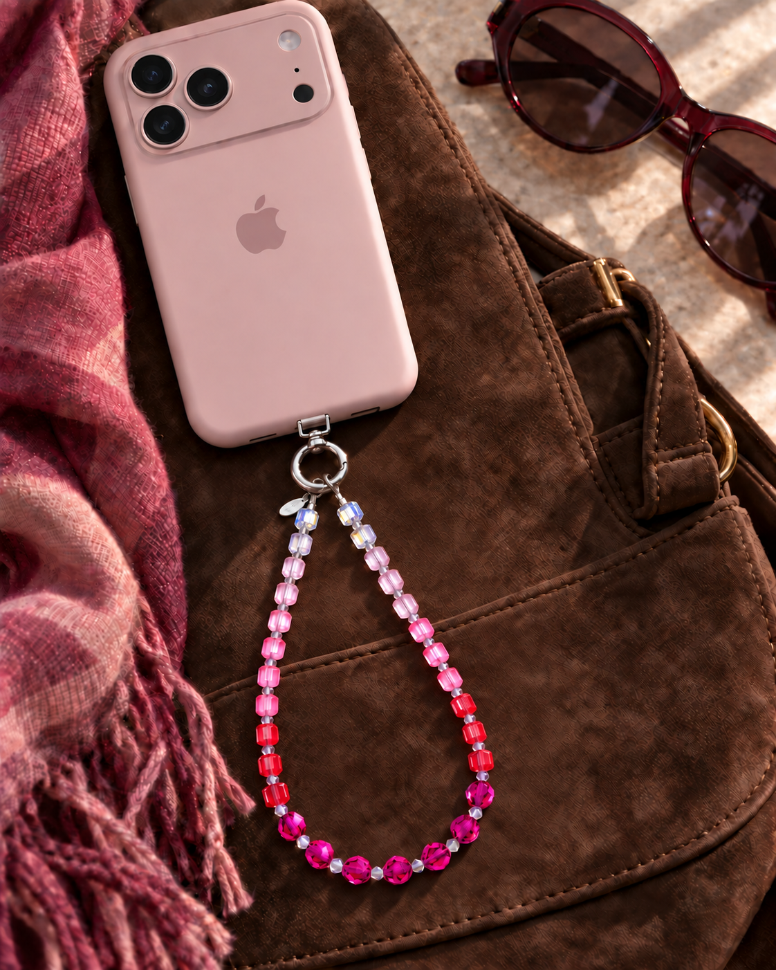 Rose ombre crystal phone chain with pale pink, pink, red and plum beads, attached to a pink phone on a brown bag.