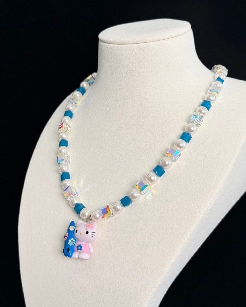 Angled view of a blue crystal and pearl necklace with a colorful cat charm on a white jewelry bust against a black background.